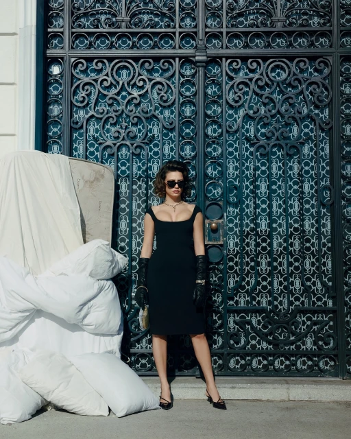 A woman poses in a black dress, gloves, and heels, against an ornamental black gate with a pile of white fabric or bedding beside her.