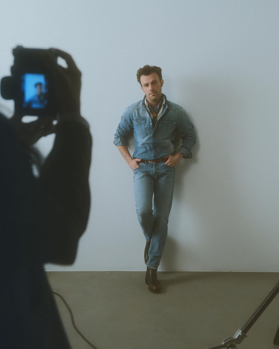 Male fashion model poses in front of a white wall while a photographer takes his picture in a light-filled studio setting.