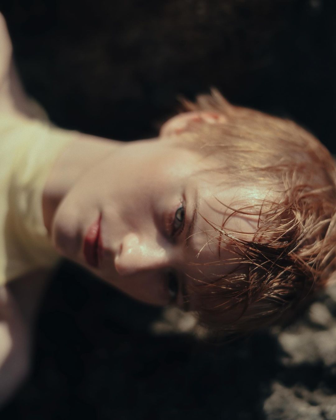 Close-up of a fair-skinned young adult with short, ginger hair and light eyes, lying down with their head tilted back on dark rocks.