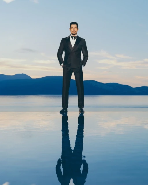 Adult male in a tuxedo standing on a reflective surface, with a lake and mountains in the background under a cloudy sky at dusk.