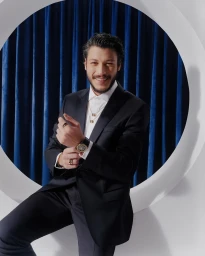 Man in a black suit adjusting his watch against a blue velvet curtain backdrop