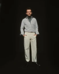 Man standing against a dark background wearing a white striped button-down shirt, grey turtleneck, and light trousers by Umur Özcan.