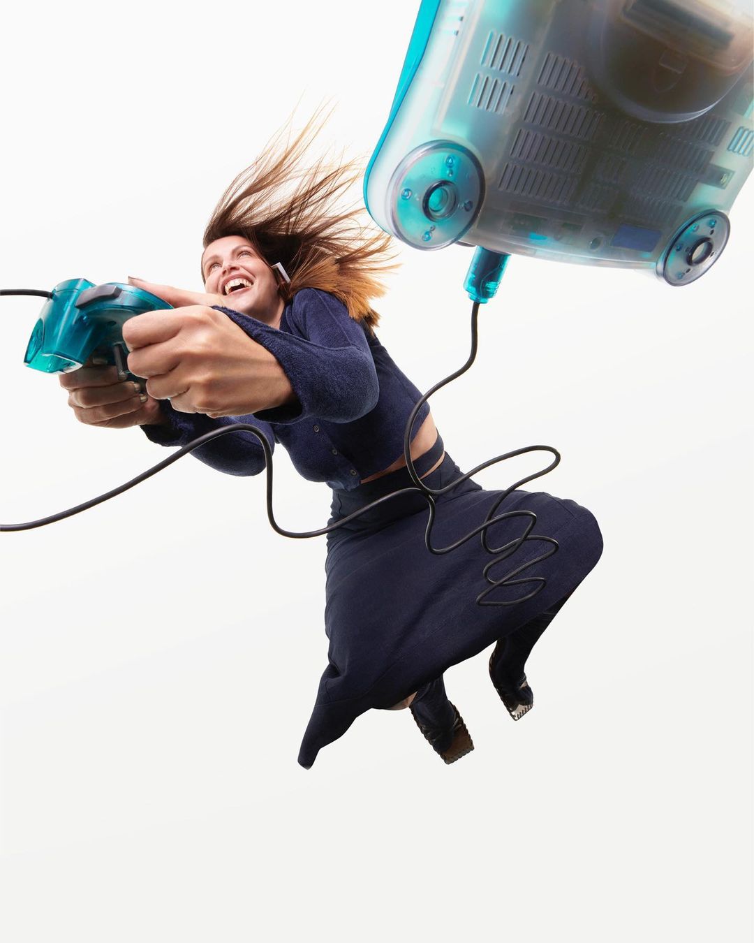 Excited woman leaps while gaming with a vintage translucent teal gamepad and connected computer in a minimalist studio, hair flying.