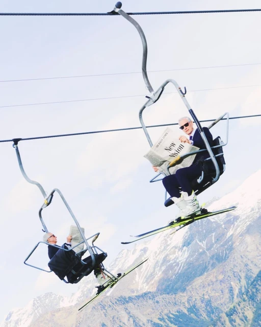 Two men ride a ski lift over a snowy mountain range, one reading a newspaper with the headline New Vision, blue sky above.
