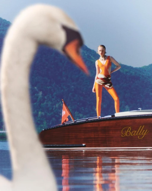A woman in orange sportswear poses on a wooden motorboat with the word 'Bally' on its side, with a swan in the foreground.