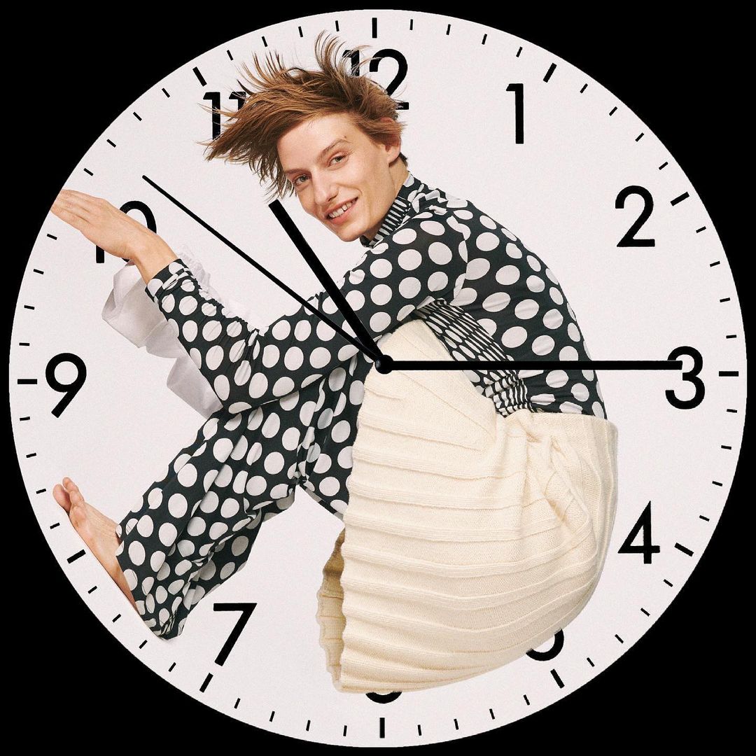 Man in polka dot top poses behind a clock face. White background, clock hands, and numbers. A fashion-forward, surreal composition.