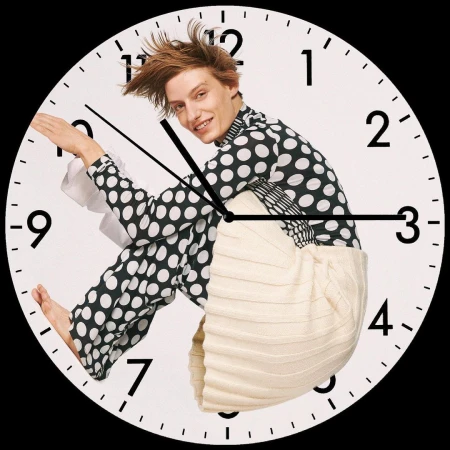 Man in polka dot top poses behind a clock face. White background, clock hands, and numbers. A fashion-forward, surreal composition.
