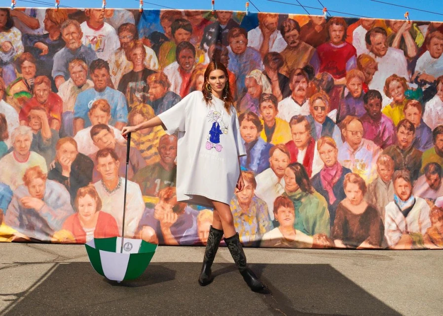 Adult female posing outdoors in front of crowd painting, wearing a white dress, black boots, and holding a green-and-white umbrella.