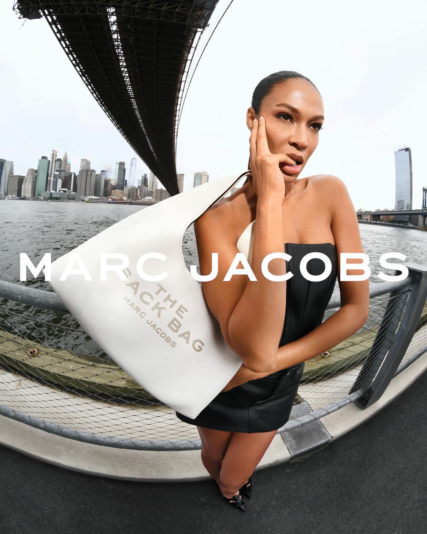 A woman in a black dress poses with a Marc Jacobs The Sack Bag against a cityscape backdrop with a bridge. Fashion editorial.