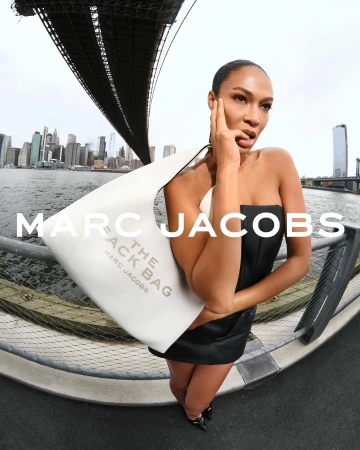 A woman in a black dress poses with a Marc Jacobs The Sack Bag against a cityscape backdrop with a bridge. Fashion editorial.