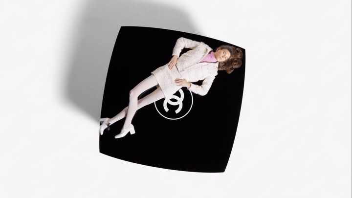 Overhead fashion shot of a woman lying on a black Chanel compact case wearing a white suit and white tights in a studio.