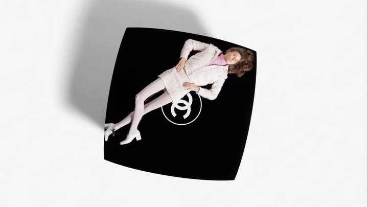 Overhead fashion shot of a woman lying on a black Chanel compact case wearing a white suit and white tights in a studio.