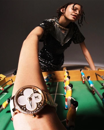 Woman in sequined dress poses with gold Louis Vuitton watch, foosball table. Fashion shot with a gray background and indoor lighting.