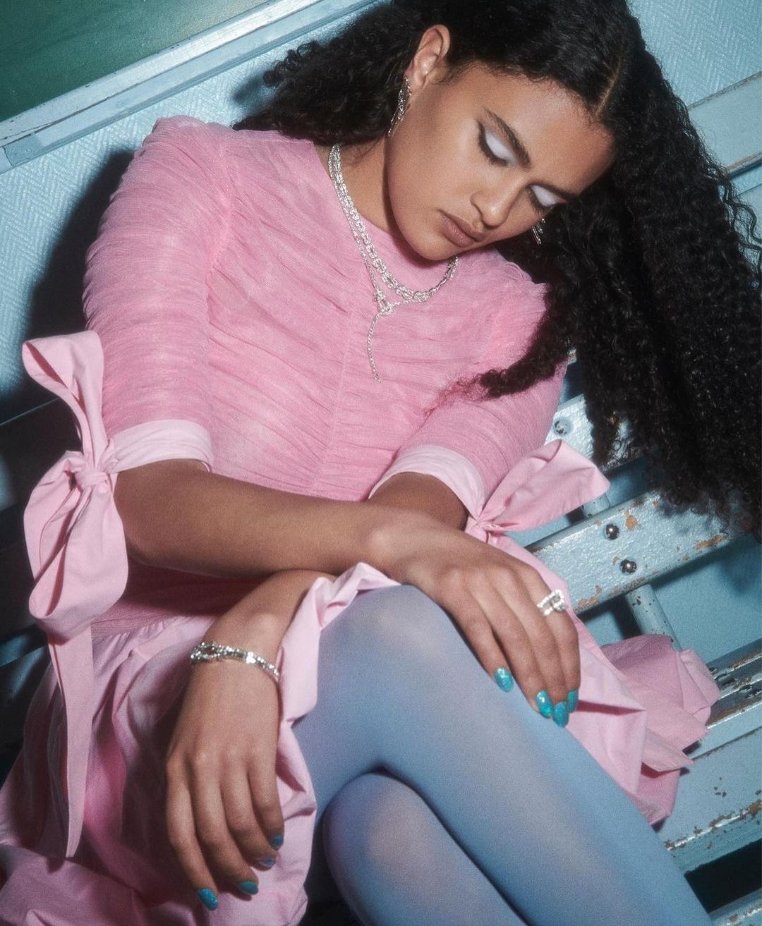 Woman in pink dress and blue tights poses with jewelry in an indoor setting, capturing fashion and beauty elements with elegance.
