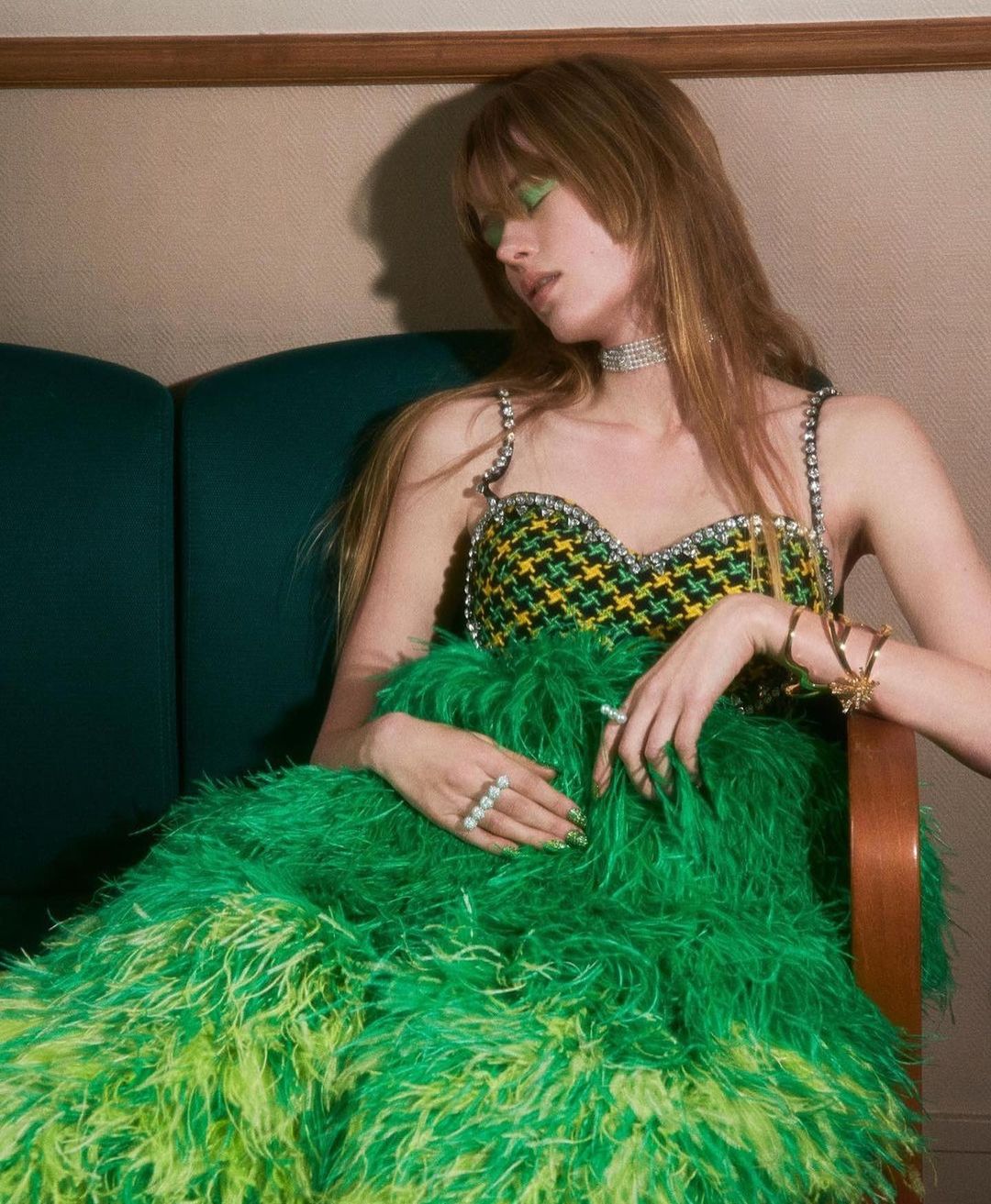 A young adult woman poses in a green and yellow feathered dress with sparkling straps, against a beige and green background indoors.