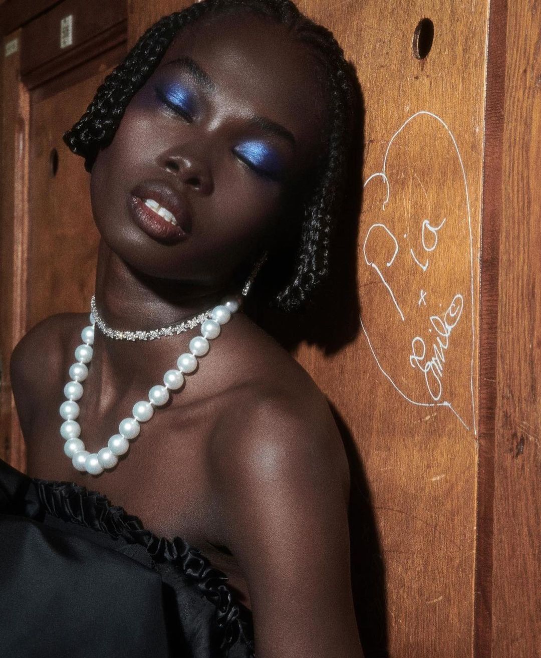 Portrait of Black female model with braided hair, wearing a strapless black dress and pearls, leaning against a wooden panel with writing.