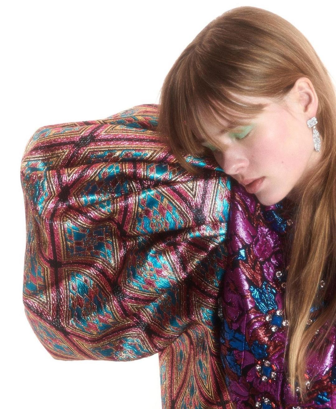 Close-up of a young woman with light brown hair resting her head on a patterned, shimmering jacket in a bright, white setting.