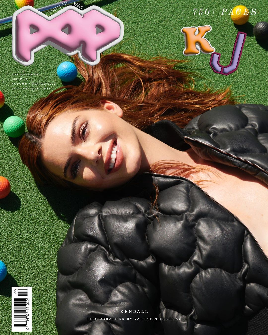 A young woman with red hair smiles while lying on a grassy surface with golf balls, wearing a black puffer jacket and glossy magazine graphics.