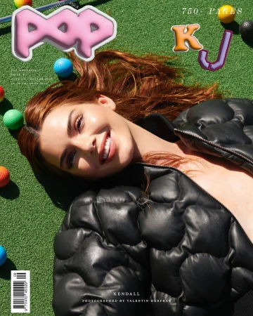 A young woman with red hair smiles while lying on a grassy surface with golf balls, wearing a black puffer jacket and glossy magazine graphics.