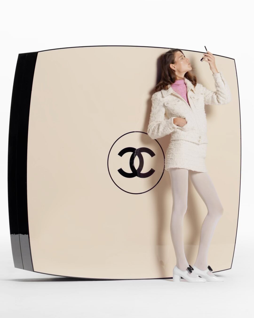 Elegant woman in white suit stands beside an oversized Chanel compact, applying makeup in a white studio, beauty and fashion shoot.