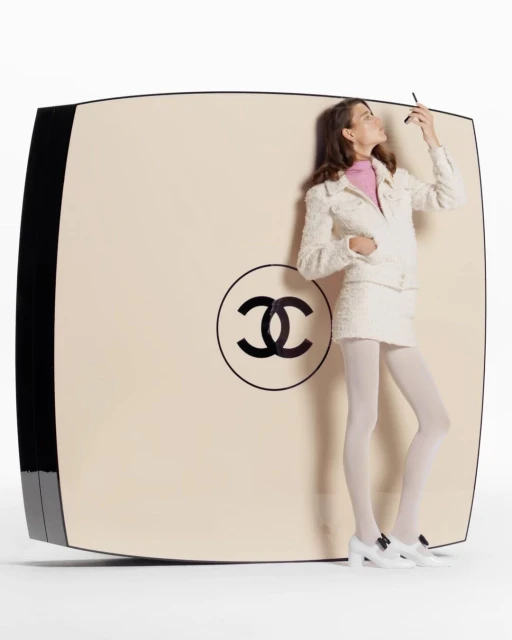 Elegant woman in white suit stands beside an oversized Chanel compact, applying makeup in a white studio, beauty and fashion shoot.