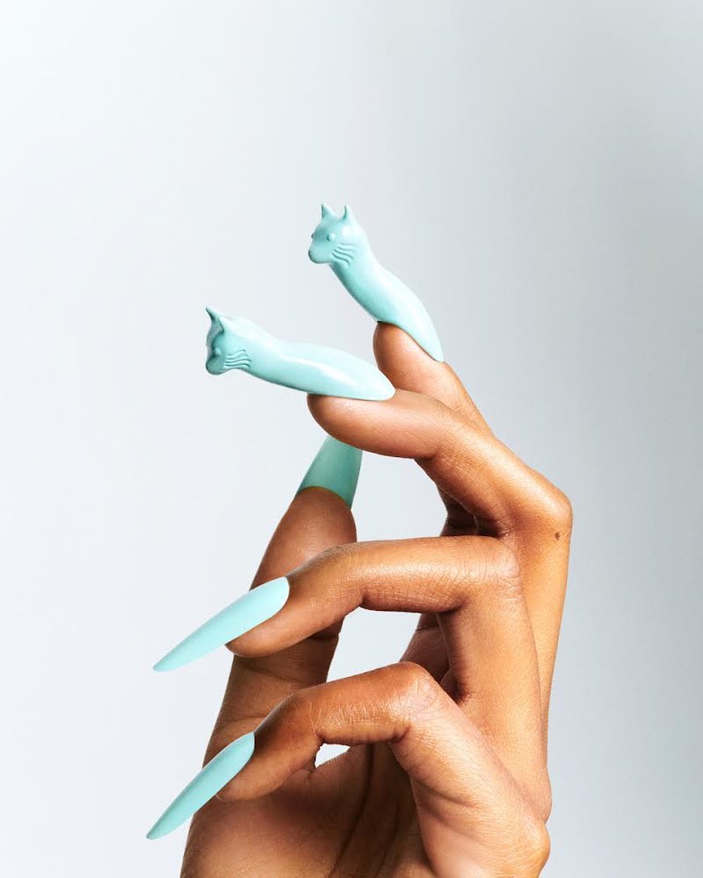 Hand with long, mint-colored stiletto nails, accessorized with sculptural cat rings, against a plain white backdrop in a studio shot.