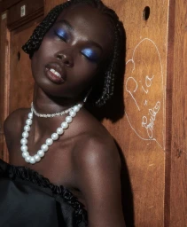 Close-up portrait of a Black woman with braided hair, striking blue eyeshadow, pearl necklace, and black top leaning against a wooden wall.