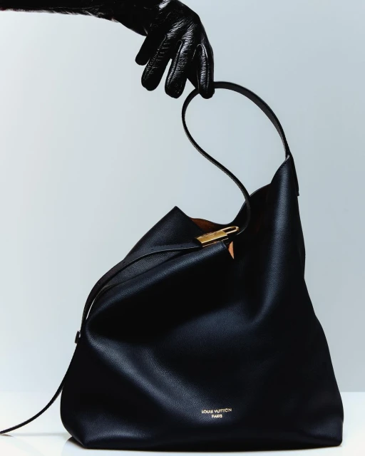 Black leather Louis Vuitton bag being held by a hand wearing a black leather glove against a light gray studio background.