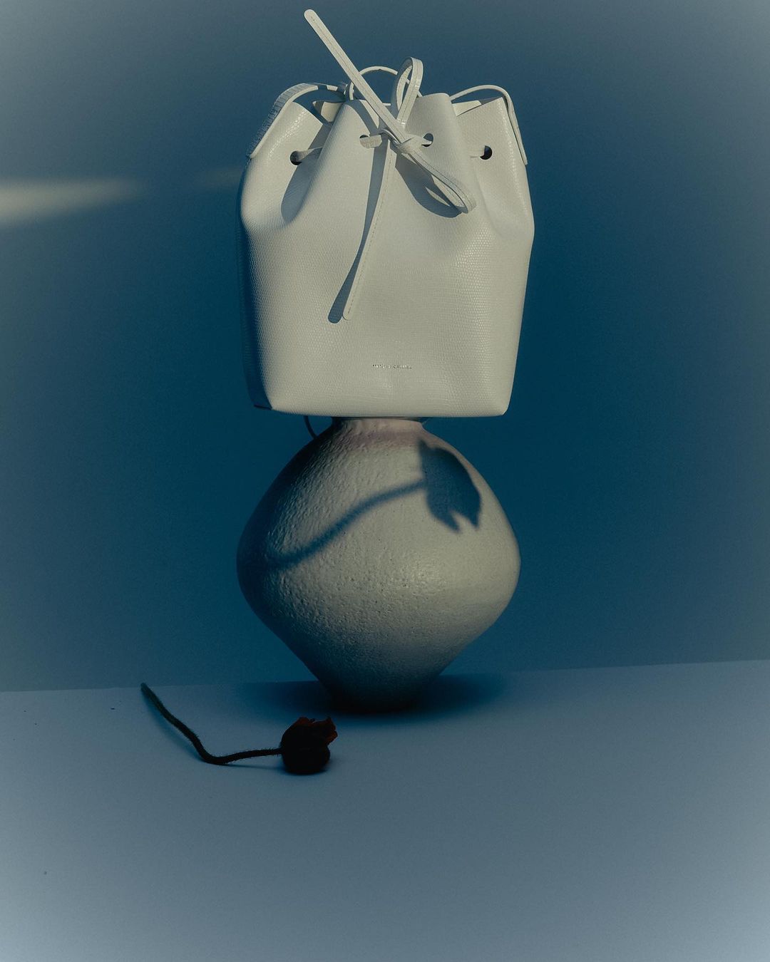 White leather bucket bag with drawstrings on a gray vase against a blue backdrop in a minimalist product shot with soft lighting.