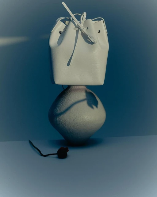 White leather bucket bag with drawstrings on a gray vase against a blue backdrop in a minimalist product shot with soft lighting.