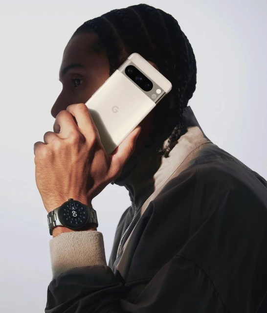Adult male in neutral jacket holds new Google Pixel phone in hand near his face, also wearing metal link watch, on light background.
