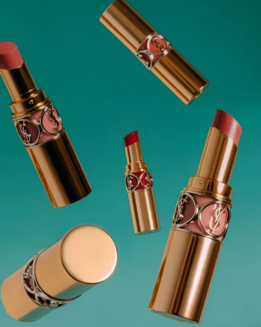 Cosmetic still life showcases five YSL lipsticks floating on a teal background. The lipsticks have gold tubes and different shades.