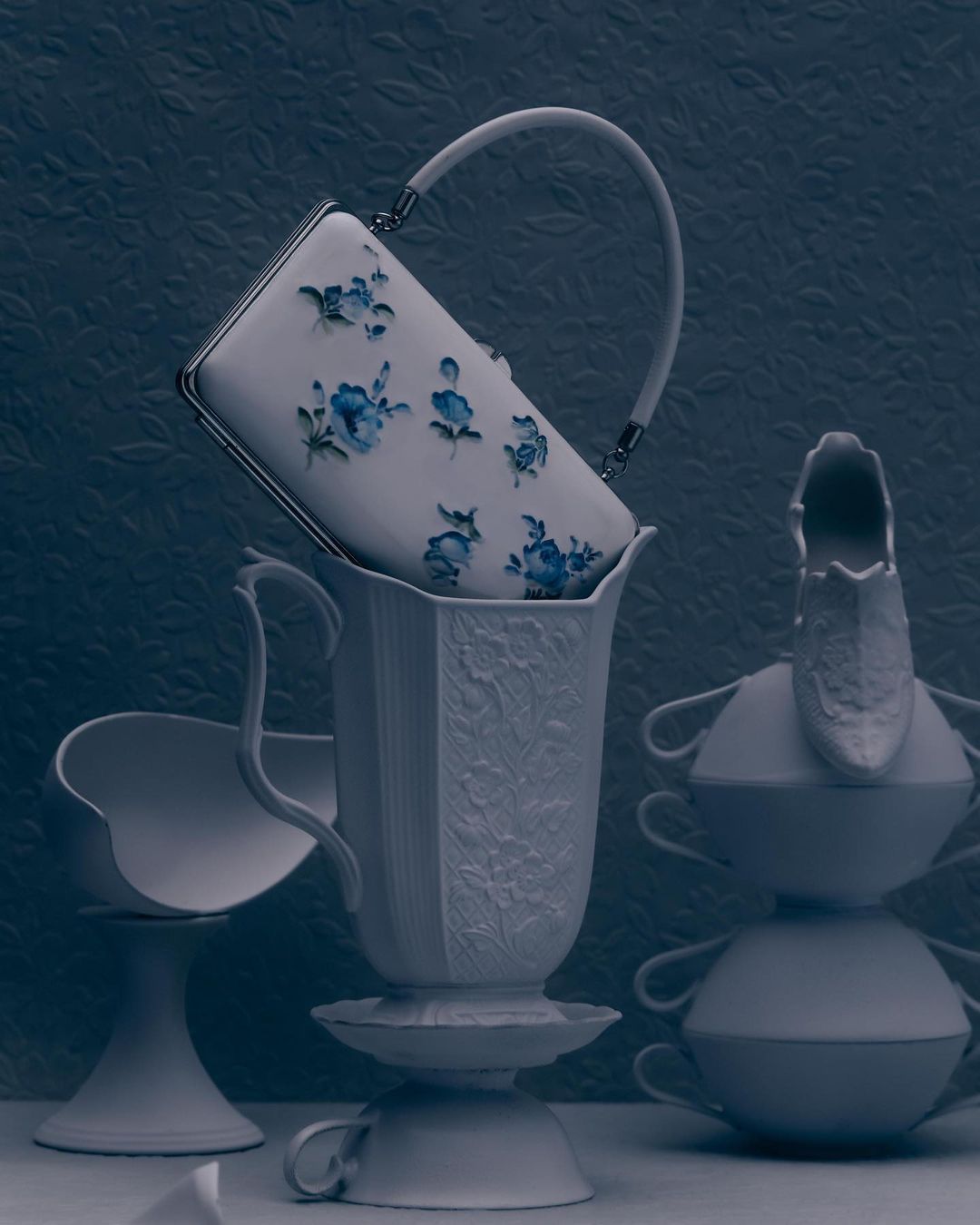 Floral handbag atop porcelain vessels, creating a whimsical still life against a textured, muted blue backdrop, showcasing fashion photography.