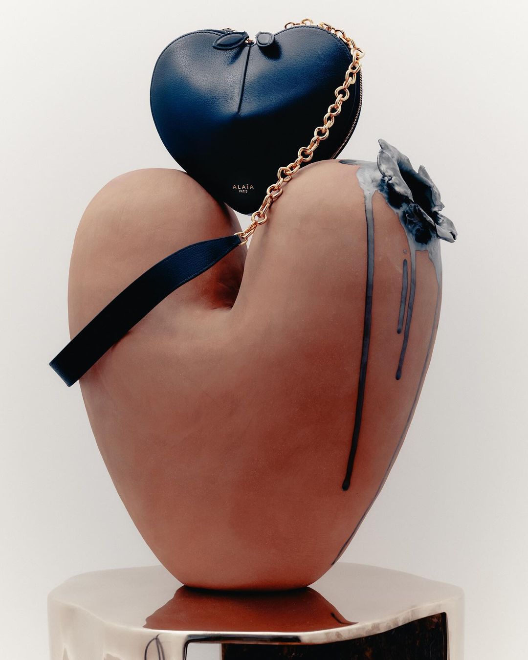 Still life featuring a heart-shaped blue purse with gold chain, displayed on a large, heart-shaped, sculpted vase with ceramic flower.