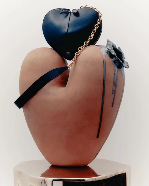 Still life featuring a heart-shaped blue purse with gold chain, displayed on a large, heart-shaped, sculpted vase with ceramic flower.