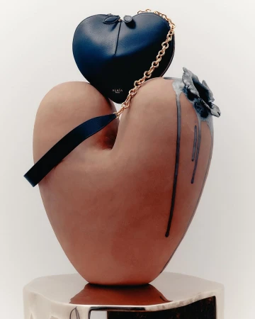 Still life featuring a heart-shaped blue purse with gold chain, displayed on a large, heart-shaped, sculpted vase with ceramic flower.