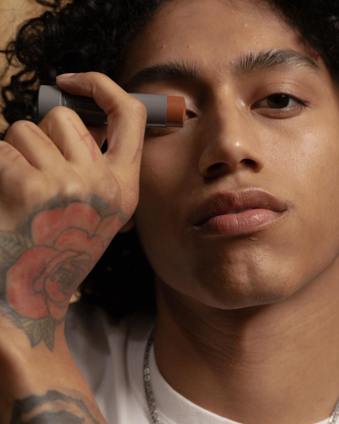 Close-up of a stylish young adult male applying face makeup. He has dark curly hair, a visible arm tattoo, and a chain necklace.