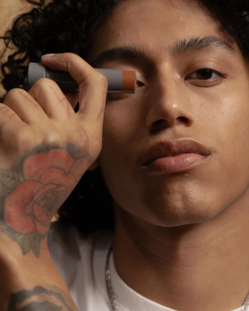Close-up of a stylish young adult male applying face makeup. He has dark curly hair, a visible arm tattoo, and a chain necklace.