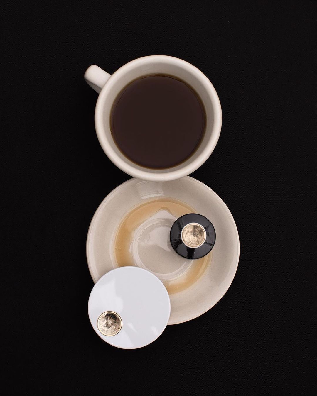 Overhead shot of a white coffee cup filled with black coffee sitting on a saucer next to two small round compact mirrors on a black background.