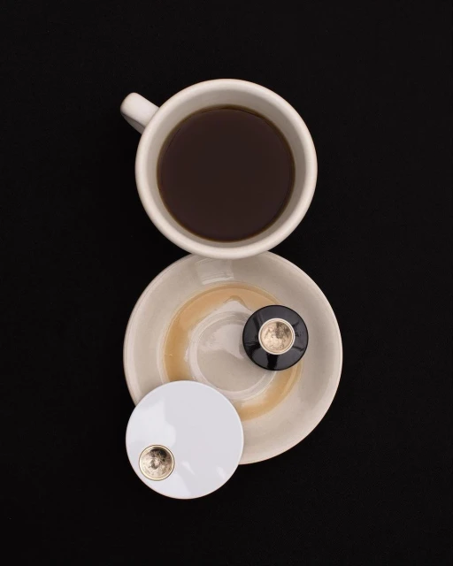 Overhead shot of a white coffee cup filled with black coffee sitting on a saucer next to two small round compact mirrors on a black background.