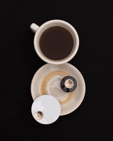 Overhead shot of a white coffee cup filled with black coffee sitting on a saucer next to two small round compact mirrors on a black background.