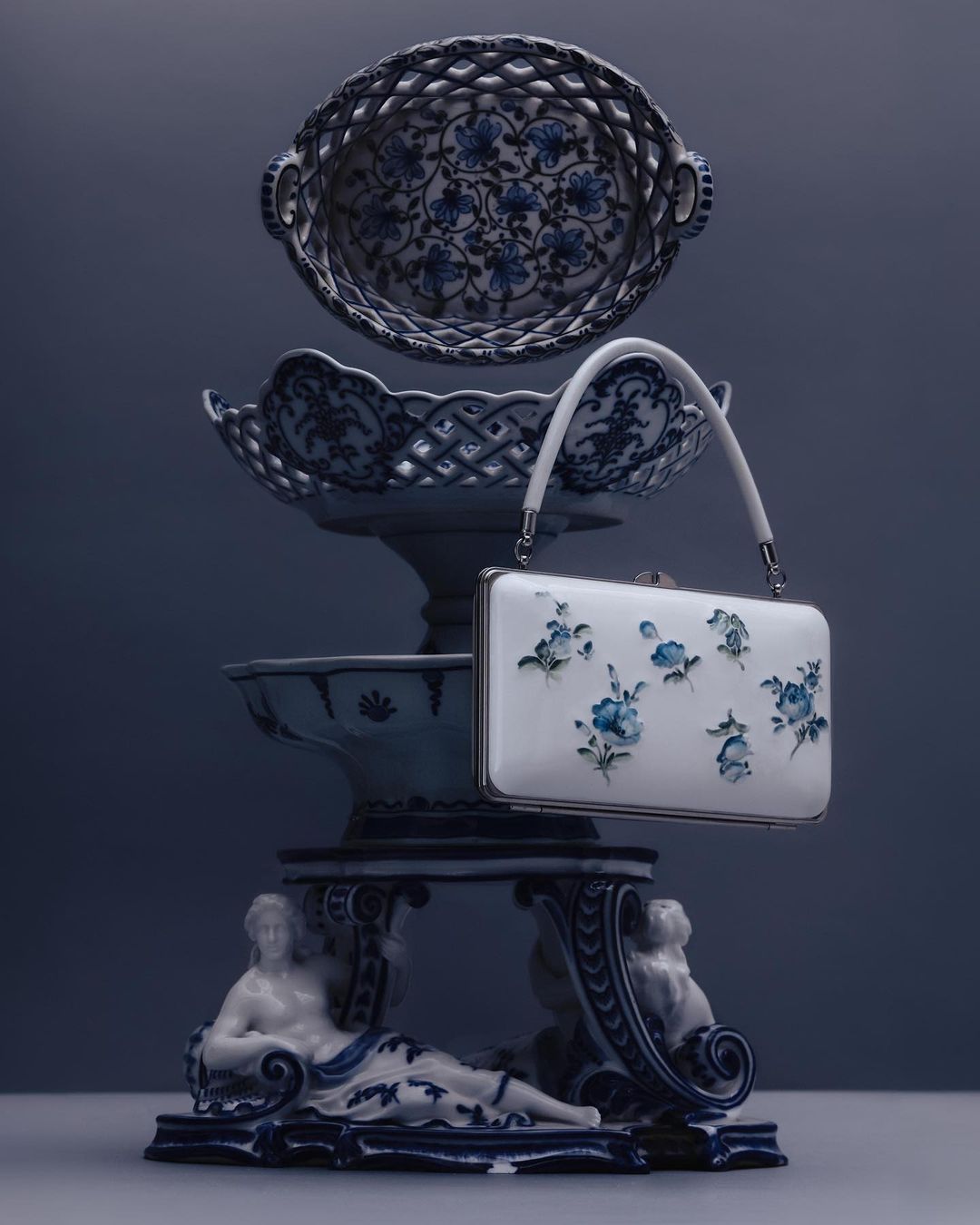 Fashion still life with white floral handbag, blue and white bowls, and porcelain figures against a blue gradient background.