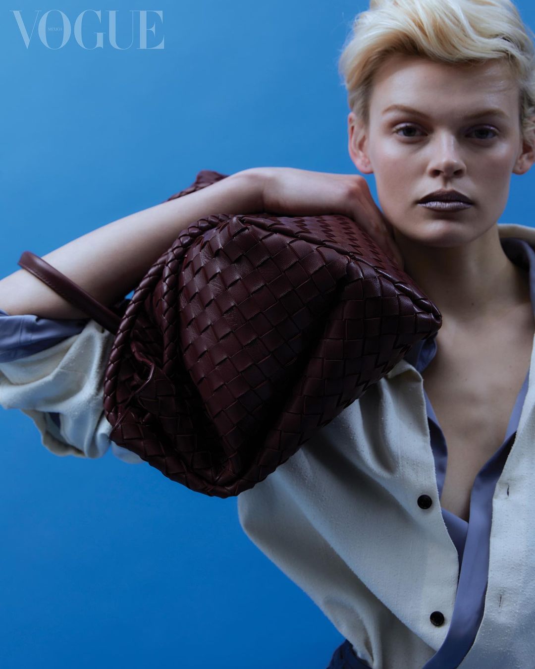 Woman with short blonde hair in studio portrait holds woven leather bag, wearing a light-colored open shirt against a blue backdrop.