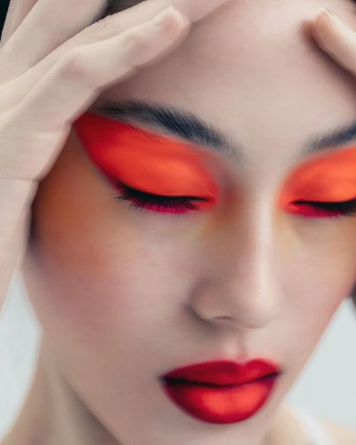 Close-up of a model wearing bold red and orange makeup and gently touching her forehead in a beauty or fashion context.