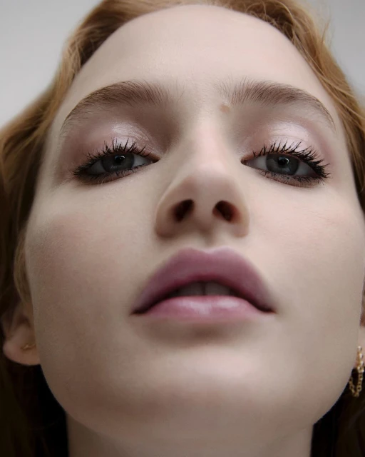 Close-up portrait of a red-haired woman with fair skin, light pink lipstick, and light eye makeup, looking up at the camera.