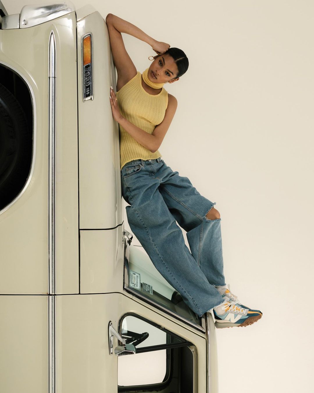A woman in casual attire poses next to a vintage vehicle with a cream-colored body, with an off-white seamless backdrop in a studio.
