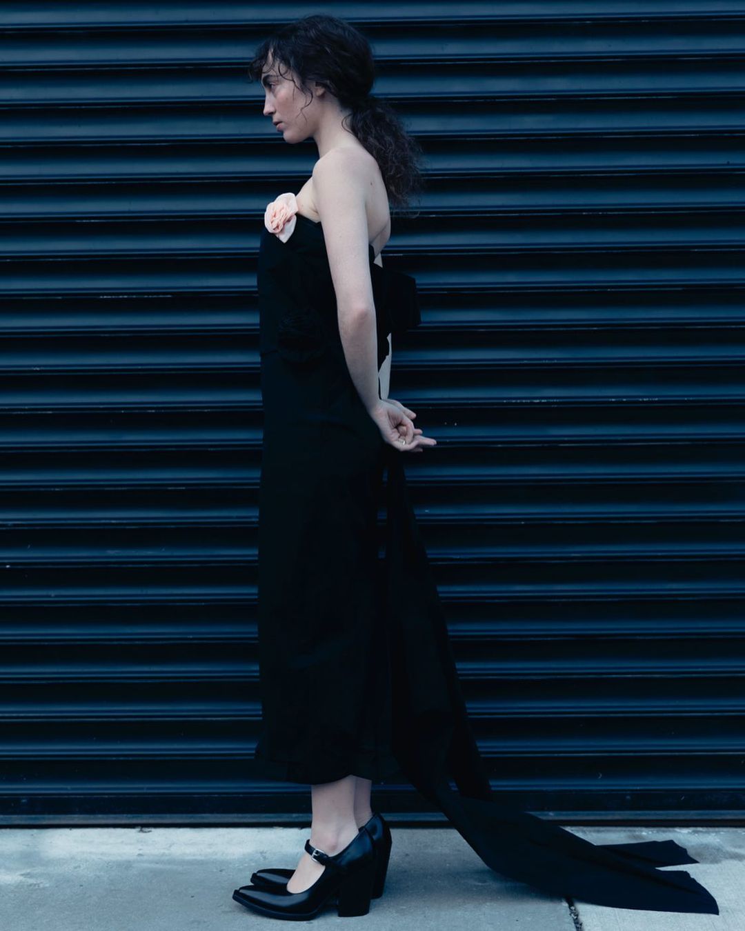 Adult woman poses in a long black dress with bow and floral accent against a dark blue corrugated metal wall. Side view, fashion shoot.