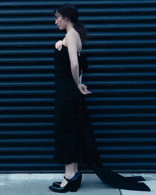 Adult woman poses in a long black dress with bow and floral accent against a dark blue corrugated metal wall. Side view, fashion shoot.