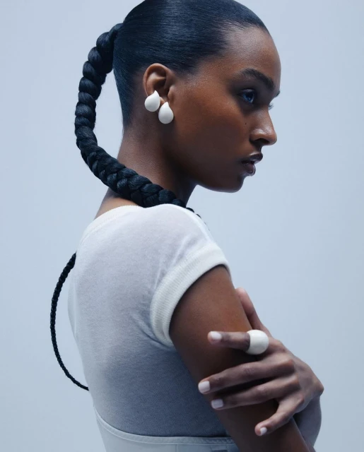 Close-up studio shot of a Black female model with a braided ponytail wearing earrings and a ring, set against a light blue background.
