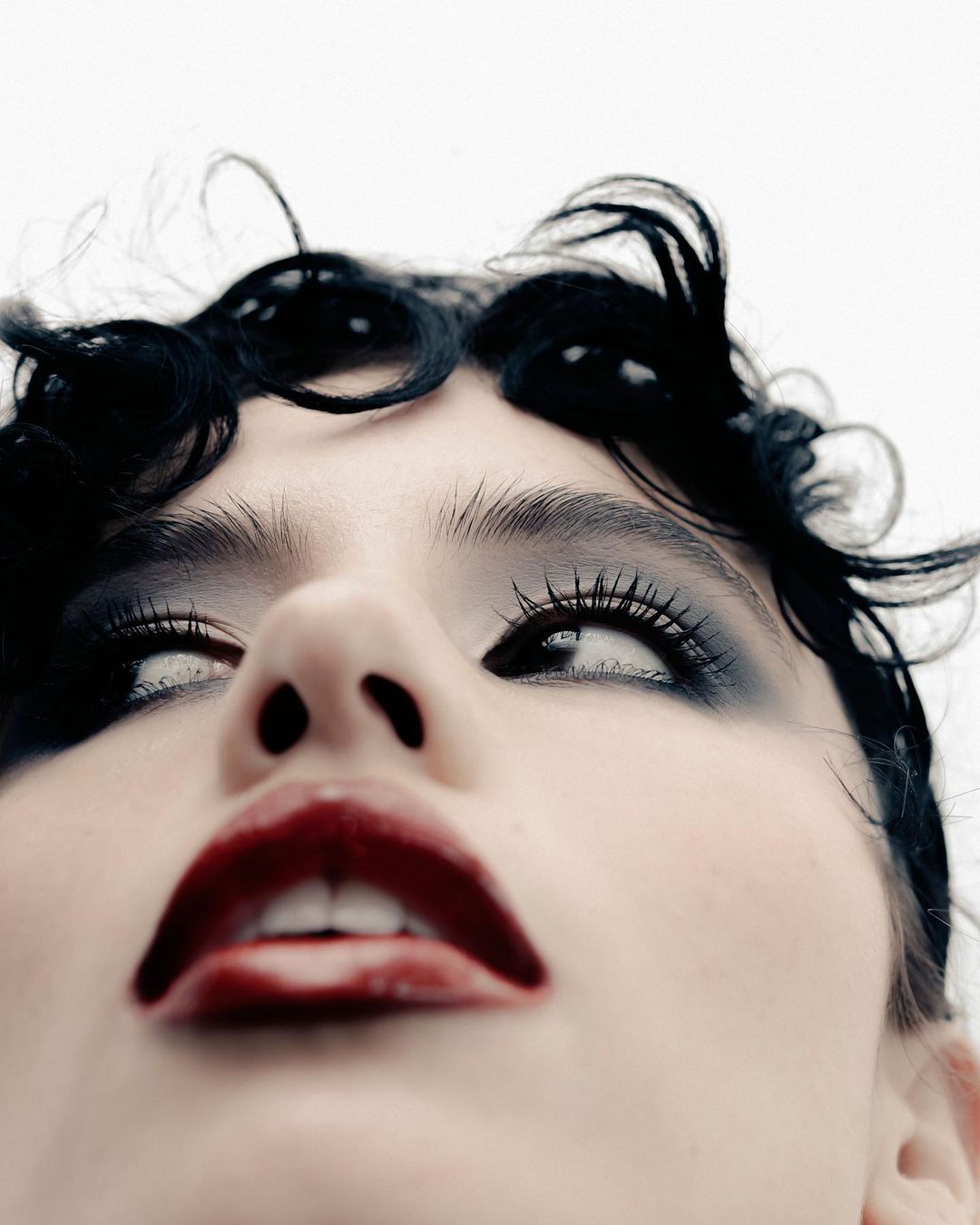 Close-up of a woman with dark hair and dramatic makeup, looking upwards, with a pale complexion and dark red lipstick.